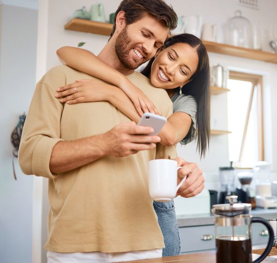 Happy young interracial couple being loving and affectionate at home. Young caucasian man using his smartphone and drinks coffee while his girlfriend embraces him from behind while they look at digital screen together