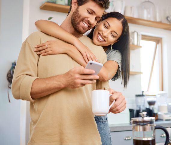 Happy young interracial couple being loving and affectionate at home. Young caucasian man using his smartphone and drinks coffee while his girlfriend embraces him from behind while they look at digital screen together