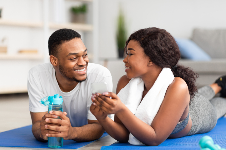 Millennial African American couple lying on sports mats with smartphone after home workout, checking new video online