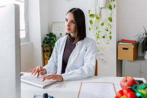 Female doctor in white coat working on laptop at office desk. Professional practitioner woman typing on keyboard looking at computer screen at clinic workplace. Healthcare people concept.