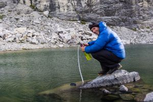 Male mountain climber in a blue down jacket filters drinking water from a lake high up in the Cordillera Blanca in the Andes in Peru