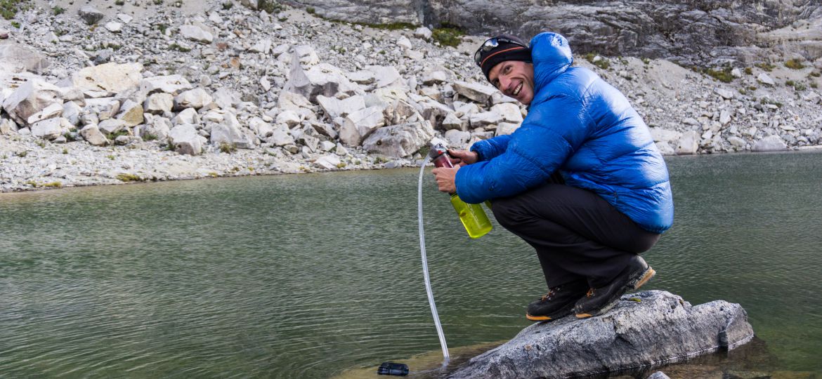 Male mountain climber in a blue down jacket filters drinking water from a lake high up in the Cordillera Blanca in the Andes in Peru