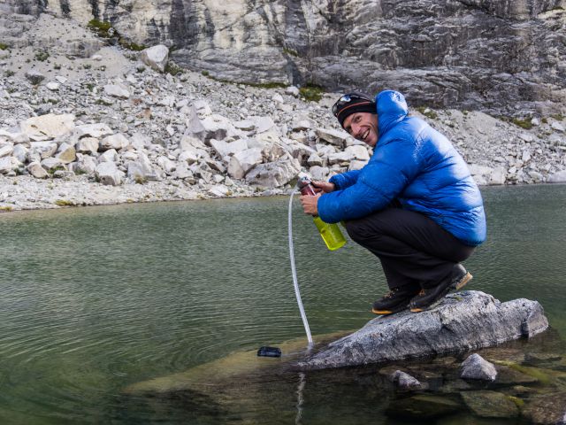 Male mountain climber in a blue down jacket filters drinking water from a lake high up in the Cordillera Blanca in the Andes in Peru